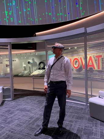 A man in formal attire stands in a modern, technology-themed space. He is wearing augmented reality glasses and is surrounded by displays and panels showcasing innovative concepts. The ceiling displays an array of digital patterns with numeric and symbolic designs.