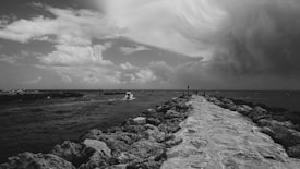 A black and white photograph captures a rocky pathway extending into the sea, with a cloudy sky overhead. The scene includes a small boat and a few figures standing along the path, suggesting a slightly dramatic and contemplative mood.