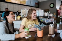 A group of three young people are sitting around a table in a café, laughing and enjoying each other's company. Two of them hold beverages in mugs with whipped cream on top. The table has small potted plants and sunglasses placed on it. The background shows a counter with menu boards and various items typically found in a café setting.