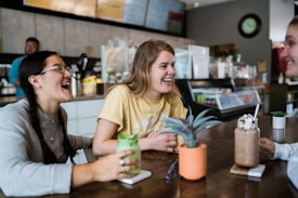 A group of three young people are sitting around a table in a caf&eacute;, laughing and enjoying each other's company. Two of them hold beverages in mugs with whipped cream on top. The table has small potted plants and sunglasses placed on it. The background shows a counter with menu boards and various items typically found in a caf&eacute; setting.