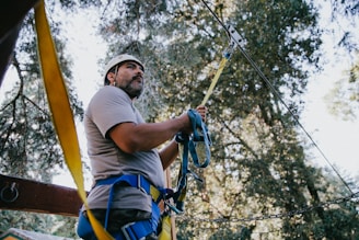 A person wearing protective gear and a helmet is standing in a forested area. They are holding onto a secured rope, possibly for a zip line or climbing activity. The background consists of tall trees and the dappled sunlight filtering through the branches.