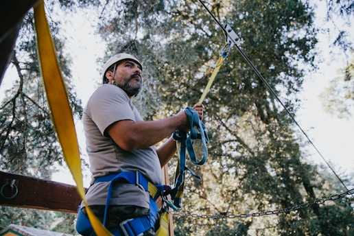 A person wearing protective gear and a helmet is standing in a forested area. They are holding onto a secured rope, possibly for a zip line or climbing activity. The background consists of tall trees and the dappled sunlight filtering through the branches.