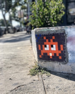 A pixel art mosaic resembling a retro video game character is embedded on a concrete surface near a sidewalk. The surrounding area includes some greenery and blurred urban elements in the background.