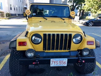 A yellow Jeep with a front view, featuring distinctive smiley face covers on its side mirrors. It is parked in an open area with some buildings and other cars visible in the background. The front bumper has additional lights and a Massachusetts license plate.
