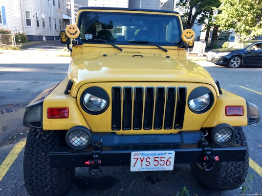 A yellow Jeep with a front view, featuring distinctive smiley face covers on its side mirrors. It is parked in an open area with some buildings and other cars visible in the background. The front bumper has additional lights and a Massachusetts license plate.