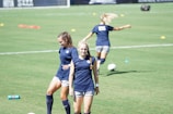 Two women are on a grassy sports field wearing athletic gear. One walks towards the camera with a smile while the other is looking down, possibly adjusting her posture. A third person is seen in the background running or stretching, partially blurred. The field is marked with cones and a soccer ball is visible.