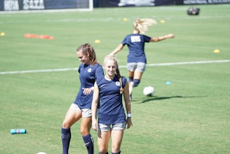 Two women are on a grassy sports field wearing athletic gear. One walks towards the camera with a smile while the other is looking down, possibly adjusting her posture. A third person is seen in the background running or stretching, partially blurred. The field is marked with cones and a soccer ball is visible.
