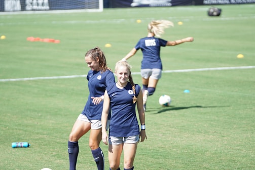 Two women are on a grassy sports field wearing athletic gear. One walks towards the camera with a smile while the other is looking down, possibly adjusting her posture. A third person is seen in the background running or stretching, partially blurred. The field is marked with cones and a soccer ball is visible.