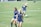 Two women are on a grassy sports field wearing athletic gear. One walks towards the camera with a smile while the other is looking down, possibly adjusting her posture. A third person is seen in the background running or stretching, partially blurred. The field is marked with cones and a soccer ball is visible.