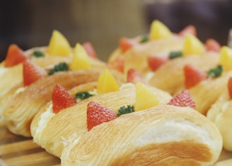 Several pastries topped with fresh strawberries, pineapple chunks, and a sprig of parsley are arranged in rows. The pastries have a flaky, golden-brown crust and a creamy filling visible on the sides.