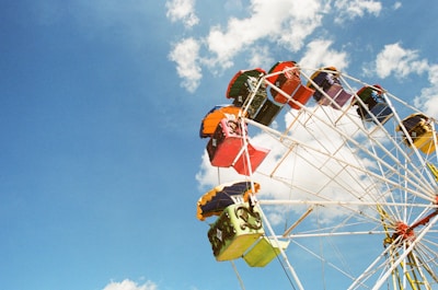 A vibrant Ferris wheel occupies the right side of the image, set against a bright blue sky with scattered white clouds. The wheel features colorful cabins in shades of red, orange, green, and yellow, creating a lively and dynamic scene.