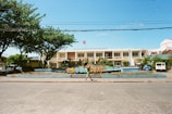 A two-story building with an architectural style characterized by large windows and a symmetrical facade, situated behind a colorful sign that spells out 'LAGONOY' amidst landscaped grounds. The scene is framed by trees on either side, and a vehicle is parked to the right. A person walks across the foreground along a paved road under a clear blue sky.