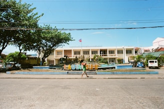 A two-story building with an architectural style characterized by large windows and a symmetrical facade, situated behind a colorful sign that spells out 'LAGONOY' amidst landscaped grounds. The scene is framed by trees on either side, and a vehicle is parked to the right. A person walks across the foreground along a paved road under a clear blue sky.