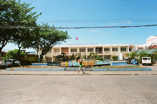 A two-story building with an architectural style characterized by large windows and a symmetrical facade, situated behind a colorful sign that spells out 'LAGONOY' amidst landscaped grounds. The scene is framed by trees on either side, and a vehicle is parked to the right. A person walks across the foreground along a paved road under a clear blue sky.