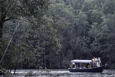 A small boat with a canopy is carrying several passengers on a calm body of water, surrounded by dense, lush green forest. The trees are tall and abundant, and some of their branches hang over the water. People on the boat appear to be engaging in recreational activities.