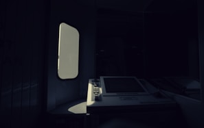A dimly lit interior with a vintage ATM or ticket machine partially illuminated by light entering through a nearby window. The shadows create a stark contrast between the bright screen and the surrounding darkness.