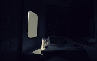 A dimly lit interior with a vintage ATM or ticket machine partially illuminated by light entering through a nearby window. The shadows create a stark contrast between the bright screen and the surrounding darkness.