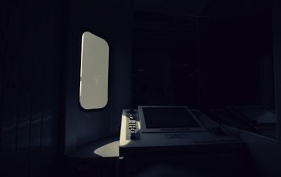 A dimly lit interior with a vintage ATM or ticket machine partially illuminated by light entering through a nearby window. The shadows create a stark contrast between the bright screen and the surrounding darkness.