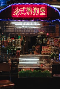 A small food stall is illuminated with a bright neon sign in Chinese characters. The counter is filled with packaged goods and fresh produce, and a woman stands behind the counter focused on her work. Various cans, bottles, and containers are displayed prominently.
