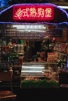 A small food stall is illuminated with a bright neon sign in Chinese characters. The counter is filled with packaged goods and fresh produce, and a woman stands behind the counter focused on her work. Various cans, bottles, and containers are displayed prominently.