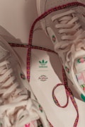 A pair of beige sneakers with colorful patterns and laces lying on a surface. The laces are multicolored with a dominant pink and green theme. The sneakers have branding marked with the 'adidas' logo and the 'AriZona' logo in green.