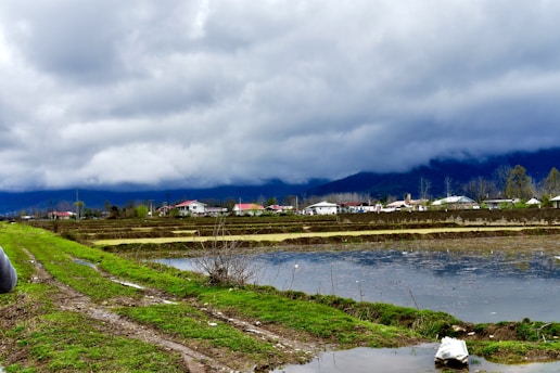 A rural landscape with vibrant green grass and muddy paths alongside water-filled fields. In the background, small houses with red and white rooftops are visible, set against a backdrop of mountains and a dramatic, cloudy sky.