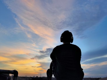 A person wearing headphones is sitting and gazing at a colorful sky filled with clouds during sunset or sunrise. The silhouette is set against a gradient of warm and cool tones, with some distant structures visible on the horizon.