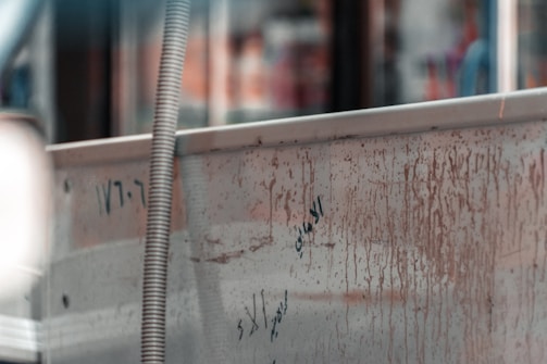 A metal surface with rust and dark marks, displaying some written numbers and letters in a possibly foreign script. A flexible hose or conduit is attached to the side, and the background is blurred with assorted colors and shapes.