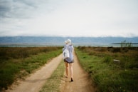 A person is walking down a dirt path bordered by green grass and shrubs. The individual is wearing a hat and casual clothing, and carrying a bag with a cross symbol. The path leads towards a large body of water and mountains in the background under a cloudy sky.