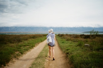 A person is walking down a dirt path bordered by green grass and shrubs. The individual is wearing a hat and casual clothing, and carrying a bag with a cross symbol. The path leads towards a large body of water and mountains in the background under a cloudy sky.