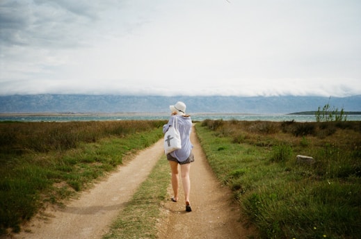 A person is walking down a dirt path bordered by green grass and shrubs. The individual is wearing a hat and casual clothing, and carrying a bag with a cross symbol. The path leads towards a large body of water and mountains in the background under a cloudy sky.