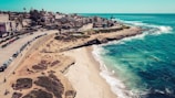 A coastal scene features a sandy beach bordered by rugged cliffs and clear blue ocean waters. The shoreline is lined with buildings and palm trees, with a road running parallel to the beach where several cars are parked. The ocean is calm, and waves gently lap against the shore.