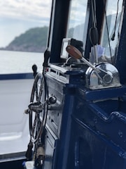 A close-up view of a ship's wheel and throttle controls inside a marine vessel, with a scenic view of the sea and distant shoreline through the window.