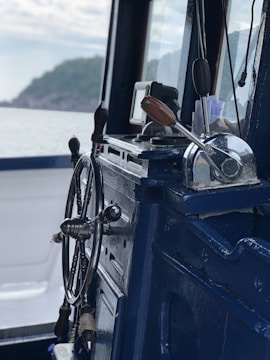 A close-up view of a ship's wheel and throttle controls inside a marine vessel, with a scenic view of the sea and distant shoreline through the window.