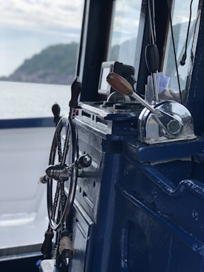 A close-up view of a ship's wheel and throttle controls inside a marine vessel, with a scenic view of the sea and distant shoreline through the window.