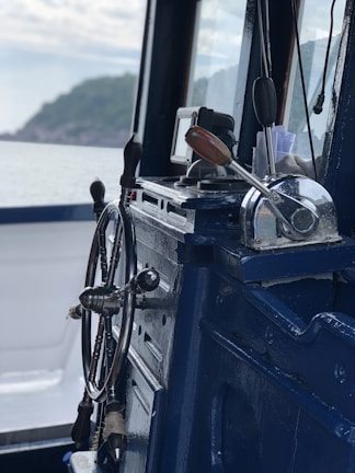 A close-up view of a ship's wheel and throttle controls inside a marine vessel, with a scenic view of the sea and distant shoreline through the window.