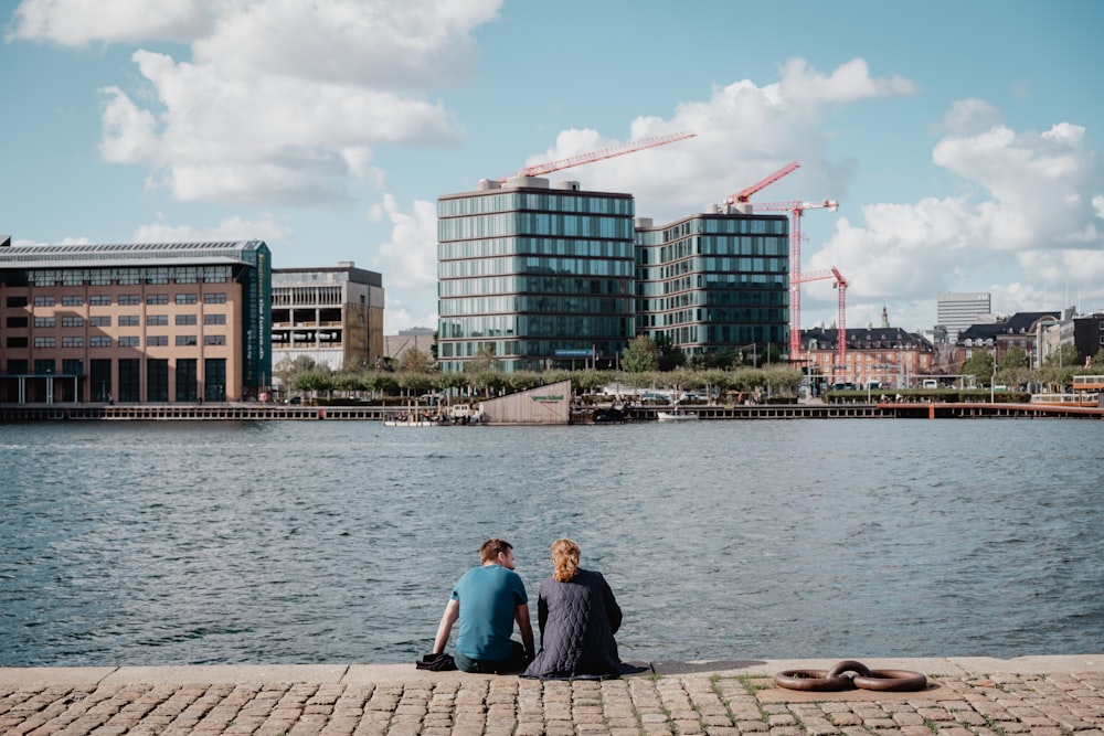 man wearing teal crew-neck shirt sitting with woman wearing gray jacket looking at lake