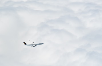 A passenger airplane is flying high above the clouds, creating a serene and peaceful scene. The aircraft is prominently featured against a backdrop of soft, fluffy clouds that stretch across the sky.