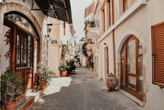 A narrow cobblestone street flanked by charming buildings with arched doorways and wooden shutters. Potted plants and small trees adorn the edges, adding greenery. The buildings have a rustic Mediterranean style, with earth tones and stone accents.