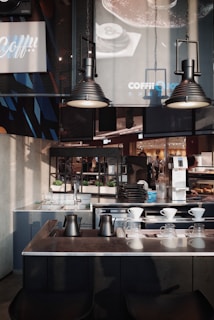 A modern coffee shop interior featuring dark metallic and minimalist design elements. The foreground shows a countertop with black metal kettles and glass drippers for pour-over coffee. Behind, there are industrial-style lamps, sinks, and an assortment of trays and coffee equipment. Posters and signs with branding details hang on the walls, alongside green potted plants placed for decoration.