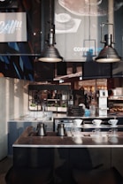A modern coffee shop interior featuring dark metallic and minimalist design elements. The foreground shows a countertop with black metal kettles and glass drippers for pour-over coffee. Behind, there are industrial-style lamps, sinks, and an assortment of trays and coffee equipment. Posters and signs with branding details hang on the walls, alongside green potted plants placed for decoration.