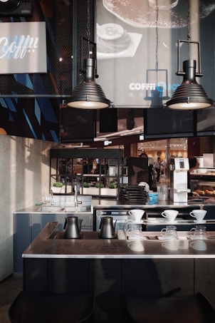 A modern coffee shop interior featuring dark metallic and minimalist design elements. The foreground shows a countertop with black metal kettles and glass drippers for pour-over coffee. Behind, there are industrial-style lamps, sinks, and an assortment of trays and coffee equipment. Posters and signs with branding details hang on the walls, alongside green potted plants placed for decoration.