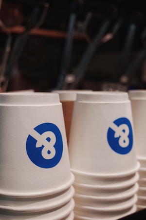 Stacks of white paper cups with a blue stylized symbol resembling the number eight. The image captures the cups prominently in focus, with blurred background elements suggesting an indoor setting.