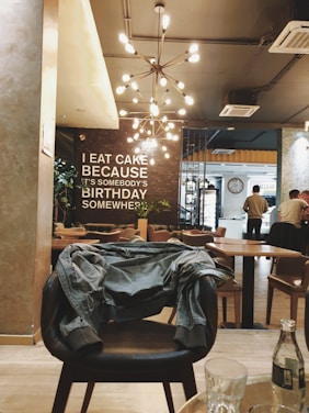A modern café interior with a quote on the wall that reads 'I eat cake because it's somebody's birthday somewhere.' The space features a stylish chandelier with multiple bulbs, warm lighting, and a mix of different seating areas, including a chair with a jacket draped over it. In the foreground, there is a small table with a bottle and glasses. Several people are seen in the background, adding a lively atmosphere.