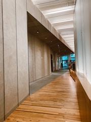 A modern corridor with wooden flooring and ceiling panels. The walls have a smooth, beige finish and are lined with recessed lights. Large windows are visible at the end, bringing natural light into the space.