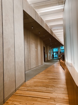 A modern corridor with wooden flooring and ceiling panels. The walls have a smooth, beige finish and are lined with recessed lights. Large windows are visible at the end, bringing natural light into the space.