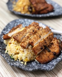 Slices of grilled chicken breast are neatly arranged on a bed of golden fried rice, all served on a rustic gray ceramic plate. The chicken is seasoned with visible herbs and spices, offering a crispy and well-cooked appearance. In the background, another plate with a similar dish is slightly out of focus.