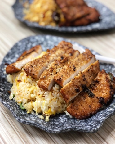 Slices of grilled chicken breast are neatly arranged on a bed of golden fried rice, all served on a rustic gray ceramic plate. The chicken is seasoned with visible herbs and spices, offering a crispy and well-cooked appearance. In the background, another plate with a similar dish is slightly out of focus.