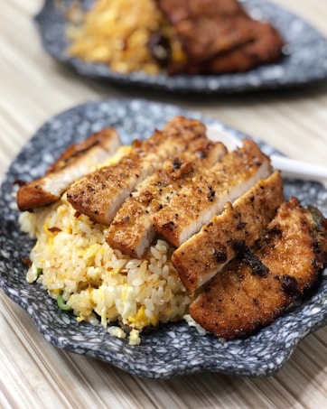 Slices of grilled chicken breast are neatly arranged on a bed of golden fried rice, all served on a rustic gray ceramic plate. The chicken is seasoned with visible herbs and spices, offering a crispy and well-cooked appearance. In the background, another plate with a similar dish is slightly out of focus.