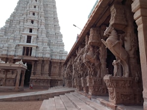 A grand temple structure showcasing intricate stone carvings and sculptures. The temple features a large towering gopuram with detailed designs, and a side view of a colonnade with horse statues on pillars. The architecture is majestic and ancient, indicative of a significant cultural and historical site.
